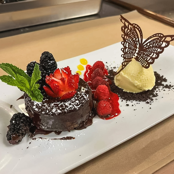 Decadent chocolate lava cake topped with a strawberry, mint, and berries, served with vanilla ice cream, raspberry sauce, and a chocolate butterfly garnish on a white plate.
