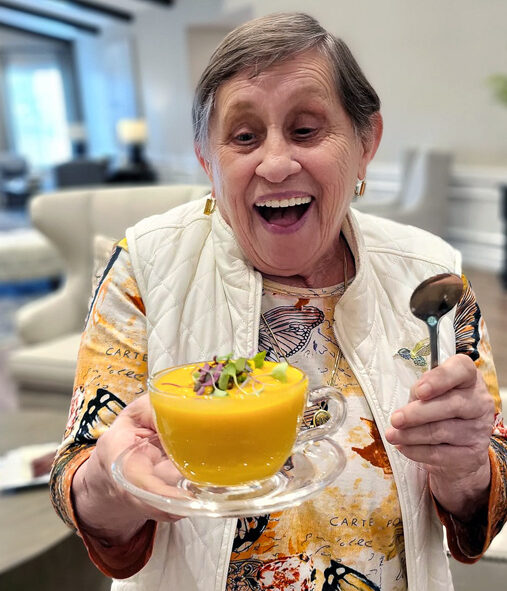 Smiling senior resident joyfully holds a bowl of vibrant orange soup garnished with greens, wearing a colorful butterfly-patterned shirt and white vest, ready to enjoy the meal.