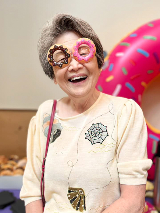 Senior resident smiles warmly while wearing a large chef's hat and a cozy sweater, creating a cheerful and festive atmosphere during a community event or celebration.