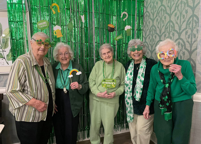 Five senior women dressed in green stand together in front of a St. Patrick’s Day-themed backdrop, smiling and holding festive props.