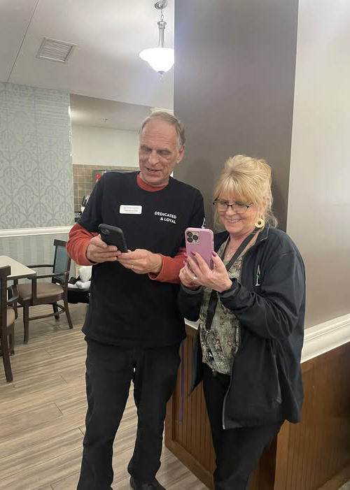 Two senior living team members stand side by side smiling, each holding a smartphone while exploring the Arrow app together in a communal space.