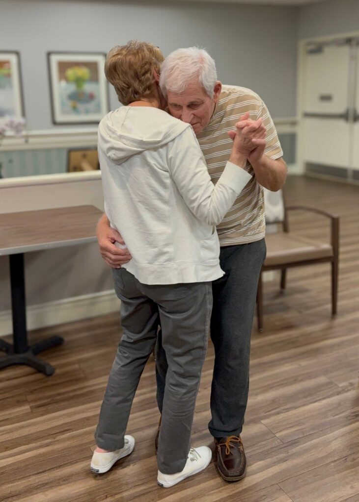 A senior couple shares a slow dance in the community room, holding each other closely and cherishing a tender moment of love and connection.