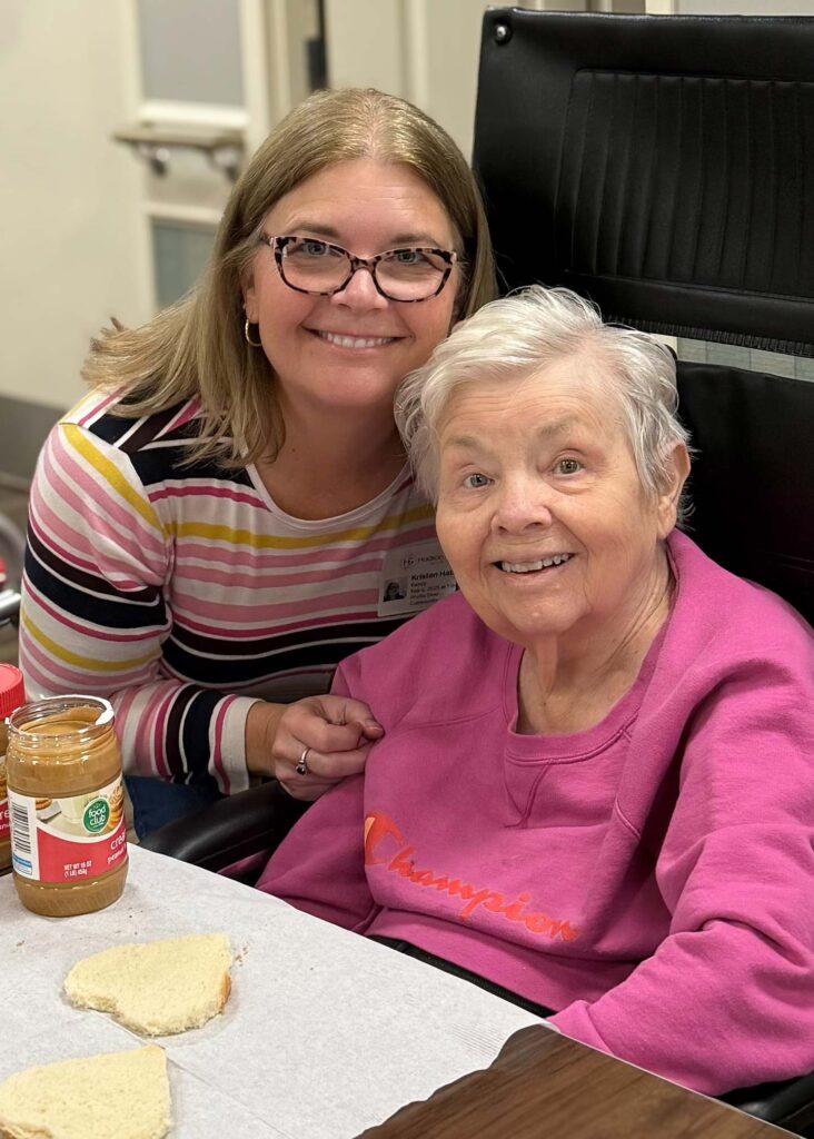 A smiling resident and team member prepare peanut butter sandwiches together at a table, enjoying a hands-on activity in the welcoming senior living community.