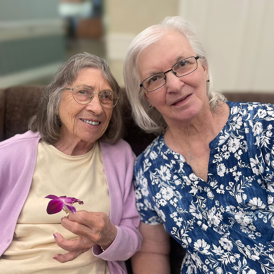 Two senior women, one holding a delicate purple flower, smile during a relaxed moment at Hudson Grande Senior Living.