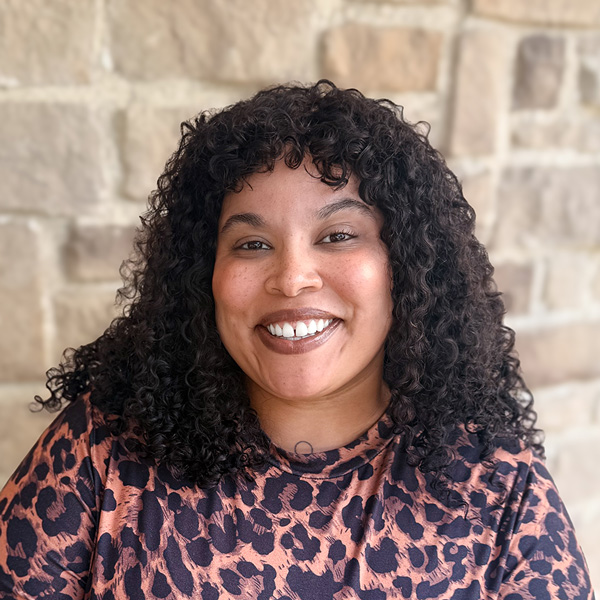 Breana Lewis, Senior Living Director at Hudson Grande Senior Living, smiling in a professional headshot with shoulder-length curly dark hair, a leopard-print blouse, and a light stone wall background.