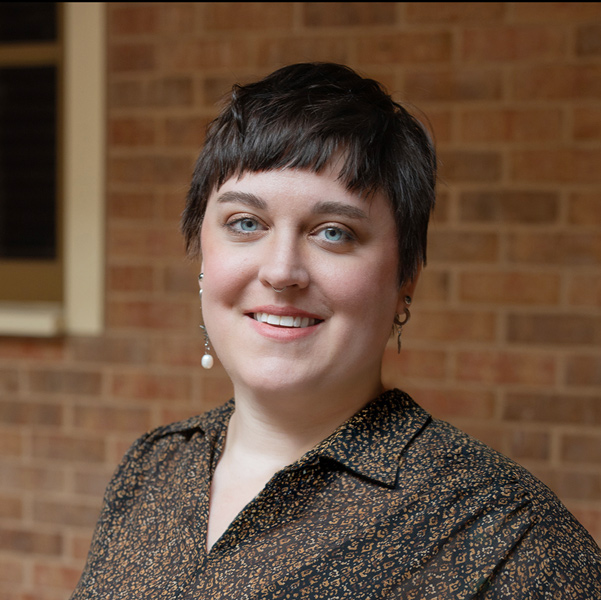 Kelsey McRill, Resident Services Director at Hudson Grande Senior Living, smiling in a professional outdoor headshot with short dark pixie-cut hair, light eyes, pearl drop earrings, a patterned blouse, and a softly blurred brick background.