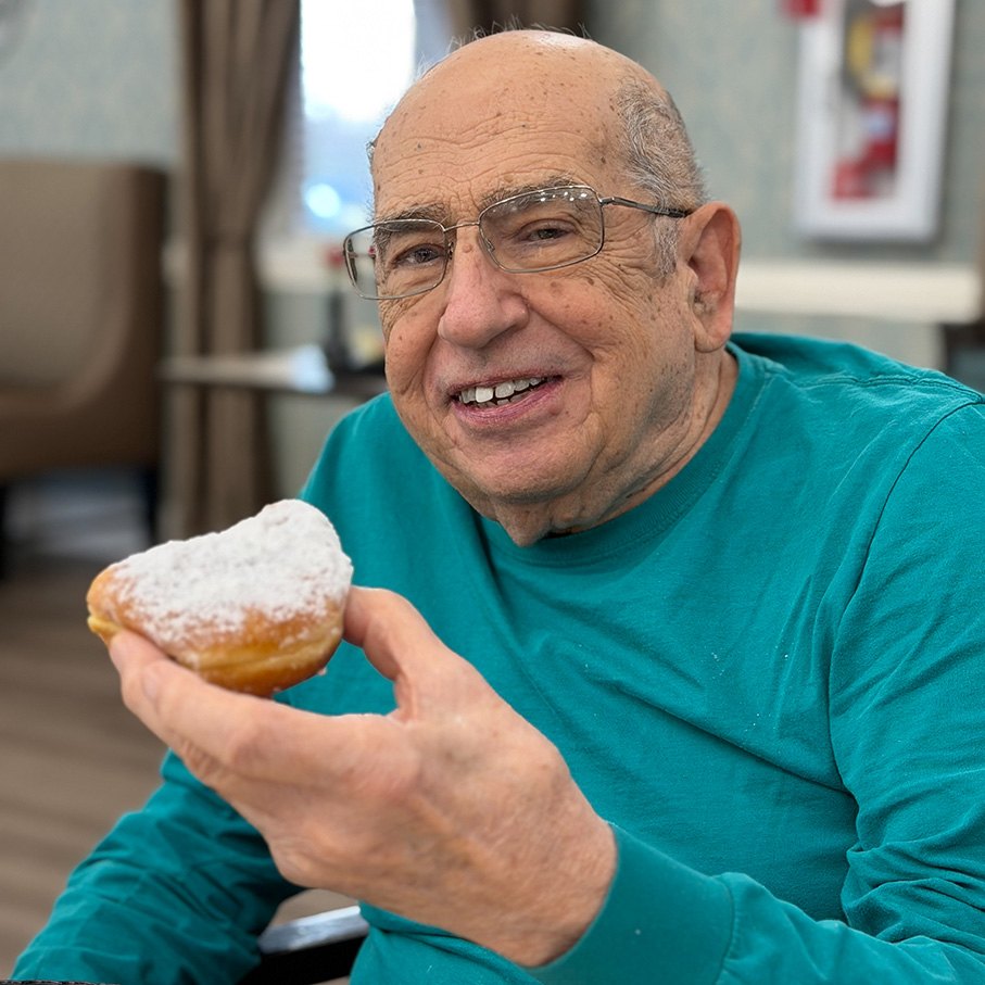 A senior man smiles, holding a donut, at Holliday Farms Senior Living.