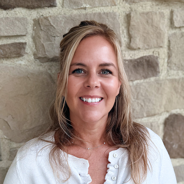 Darcy Ebie, Senior Living Coordinator at Hudson Grande Senior Living, smiling in a professional headshot with blonde hair, a white top, and a light stone wall background.